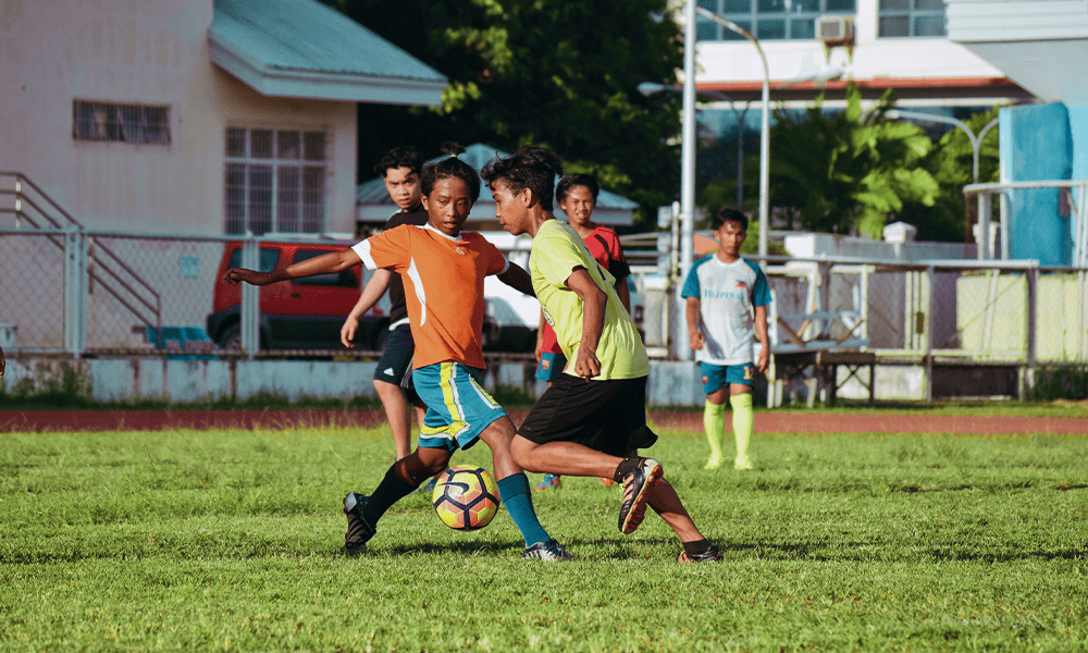 Young players in bright sports attire are playing soccer on a grass field, with a school building in the background.