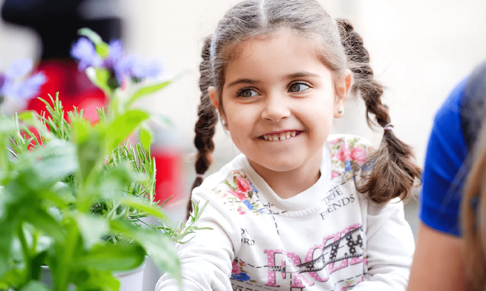 A child with a joyful expression looks at plants, wearing a floral and text-patterned top.