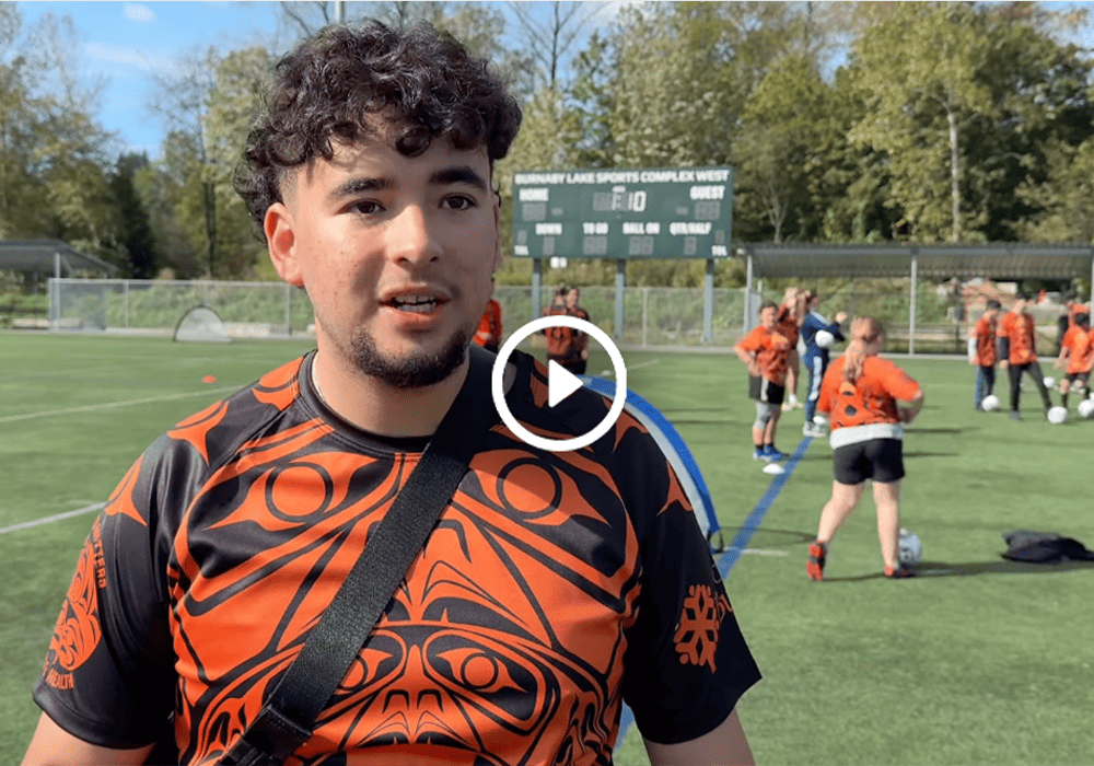 An individual in an orange and black jersey stands at a sports complex, with athletes playing in the background.