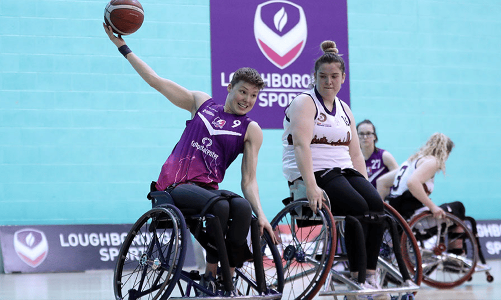 Two wheelchair basketball players in action during a game, one wearing a purple Loughborough University jersey stretching to pass the ball, while the other, in a white jersey, plays defense. They are on a court with a Loughborough Sports logo visible in the background.